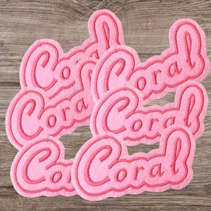May include: Several pink felt patches, each embroidered with the word "Coral" in a darker pink. The patches feature a rounded, cursive font and are arranged on a wooden surface.
