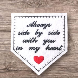 May include: White felt banner with black stitched border and text "Always side by side with you in my heart." A small red heart is stitched at the bottom. The banner is on a wooden surface.
