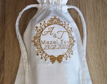 May include: White drawstring bag with gold embroidery. The embroidery features a decorative frame with the initials "A & T", the text "Mazel Tov", and the date "23.02.2022".