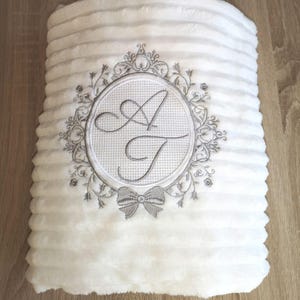May include: White plush blanket with a grey embroidered monogram of the letters "A" and "T" in a decorative frame with a bow.