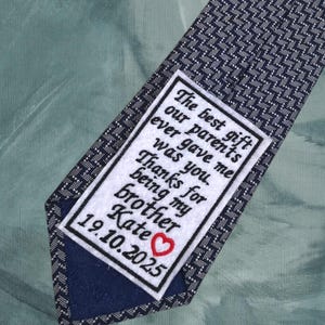 May include: Navy blue tie with a white and navy geometric pattern. A white embroidered patch features the text "The best gift our parents ever gave me was you. Thanks for being my brother Kate 19.10.2025" with a red heart.