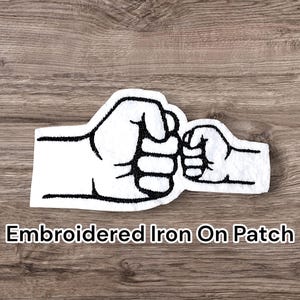 May include: Embroidered iron-on patch showing two white fists bumping, with black outlines. The patch has a felt-like texture and the text "Embroidered Iron On Patch" is displayed below the image.