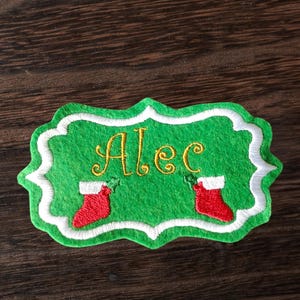 May include: Green felt Christmas decoration with the name "Alec" in gold script. The ornament has a white scalloped border and features two red stockings with white cuffs.