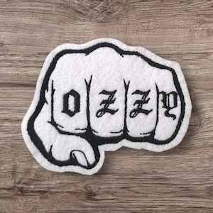May include: A white patch shaped like a clenched fist, featuring the word "OZZY" in black gothic script across the knuckles. The patch has a black outline and is set against a wooden surface.