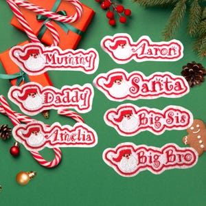 May include: Embroidered Christmas name patches featuring Santa Claus designs. Names include "Mummy", "Daddy", "Amelia", "Aaron", "Santa", "Big Sis", and "Big Bro". The patches have a red and white color scheme, perfect for holiday gifts.