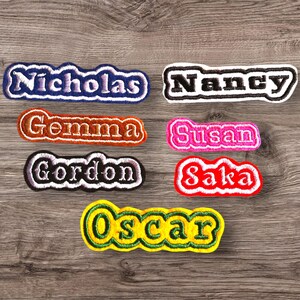 May include: Seven colorful embroidered iron-on name patches with white lettering. The names are Nicholas, Nancy, Gemma, Susan, Gordon, Saka, and Oscar.