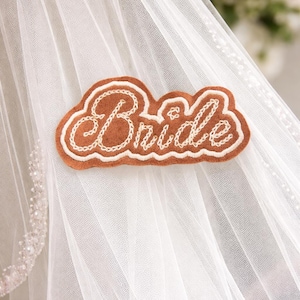 May include: A brown felt badge with the word "Bride" embroidered in white cursive lettering. The badge is in the shape of a cloud and is attached to a white tulle veil, with a beaded edge.