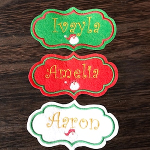 May include: Three embroidered Christmas name tags in green, red, and white. Each tag has a scalloped edge with a contrasting border. The names "Ivayla", "Amelia", and "Aaron" are embroidered in gold, with a small Santa Claus design.