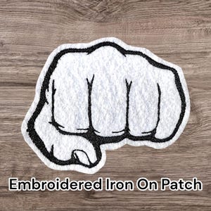 May include: White embroidered iron-on patch in the shape of a clenched fist with a black outline. The patch has the text "Embroidered Iron On Patch" in a black font.