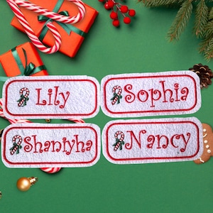 May include: Four white rectangular name tags with red trim and embroidered names: Lily, Sophia, Shaniyah, and Nancy. Each tag features a candy cane and holly design. The tags are on a green surface with Christmas decorations.