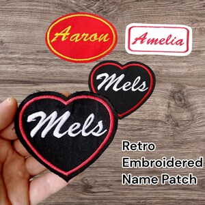 May include: Embroidered name patches in red, black, and white. The patches include the names "Aaron", "Amelia", and "Mels". The patches are in various shapes, including an oval, a rectangle, and a heart. The text "Retro Embroidered Name Patch" is also visible.