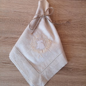 May include: A beige linen napkin with a white embroidered wreath and a bunny inside. The napkin is tied with a brown twine bow.