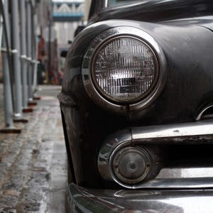 May include: Close-up of a classic black vintage car's front end. The image highlights the round headlight with a chrome bezel, a smaller turn signal light, and a shiny chrome bumper. The car is parked on a wet cobblestone street. Text: A. Hickman - 2025.