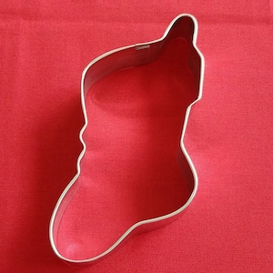 May include: A silver metal cookie cutter shaped like a stocking. The cutter is on a red background.