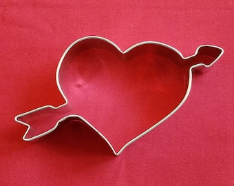 May include: A heart-shaped cookie cutter with an arrow through it, made of silver-colored metal. The cutter is on a red background, creating a Valentine's Day theme.