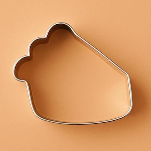 May include: A pie-shaped cookie cutter made of shiny, silver-colored metal. The cutter has a scalloped edge on one side, and a straight edge on the other. The background is a solid, light orange color.