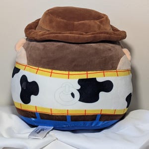 May include: A plush toy shaped like the character Woody from Toy Story. The toy has a brown cowboy hat, brown hair, and a white vest with black cow spots. The vest is trimmed with yellow and red details, and blue pants.