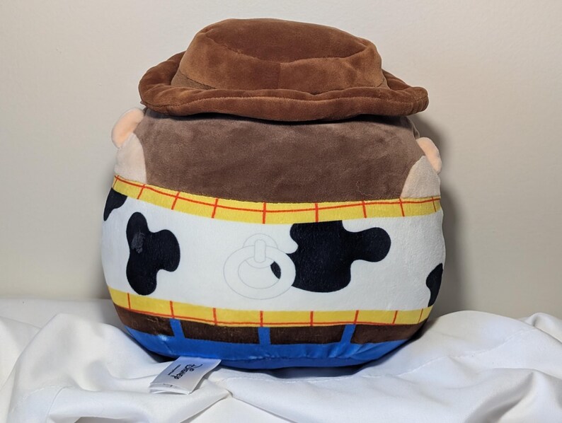 May include: A plush toy shaped like the character Woody from Toy Story. The toy has a brown cowboy hat, brown hair, and a white vest with black cow spots. The vest is trimmed with yellow and red details, and blue pants.