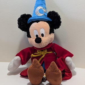 May include: A plush Mickey Mouse dressed as a sorcerer. Mickey wears a blue wizard hat with a silver crescent moon and star, a red robe, and brown shoes. His black ears and white gloves are visible.