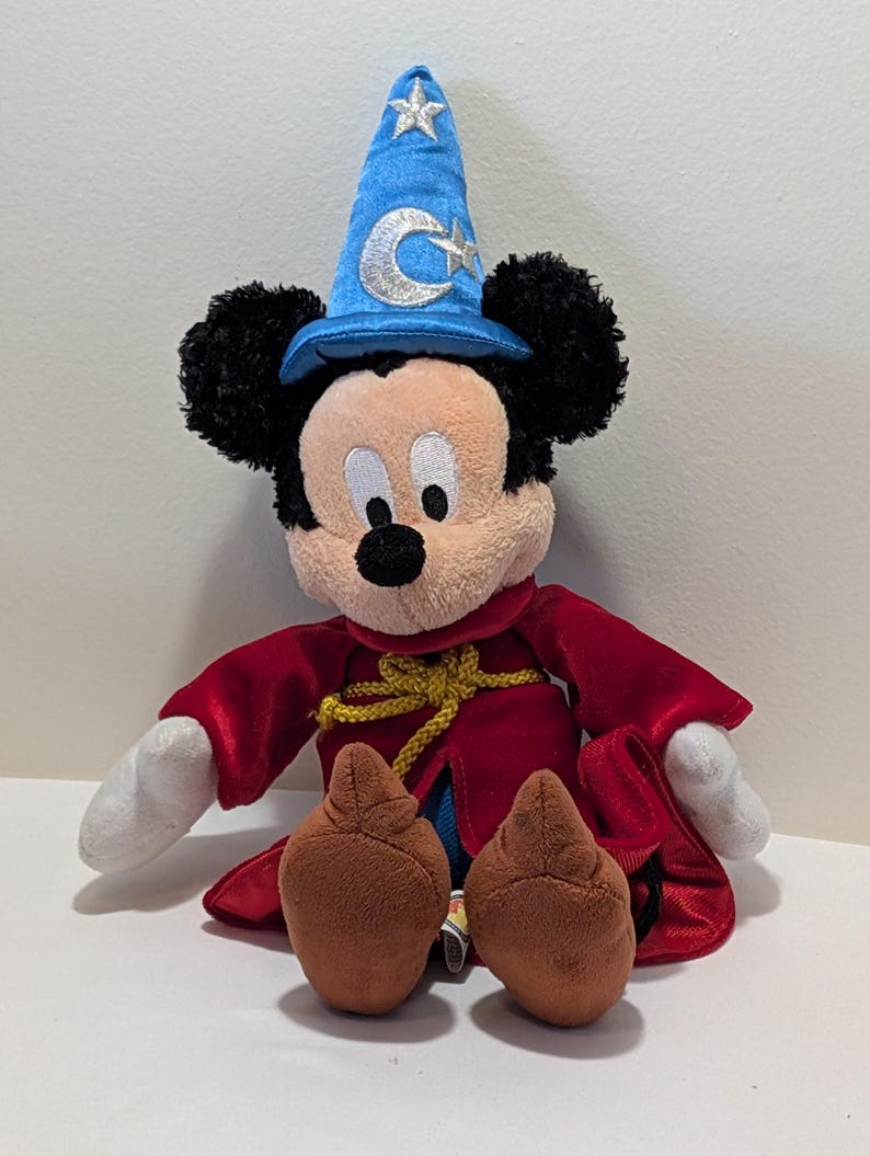 May include: A plush Mickey Mouse dressed as a sorcerer. Mickey wears a blue wizard hat with a silver crescent moon and star, a red robe, and brown shoes. His black ears and white gloves are visible.