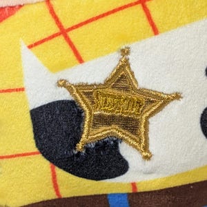 May include: Close-up of a plush toy featuring a gold sheriff's star with the word "SHERIFF" embroidered on it. The star is on a white background, with black and yellow fabric visible.