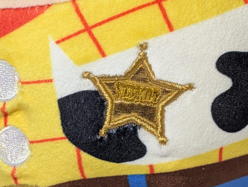 May include: Close-up of a plush toy featuring a gold sheriff's star with the word "SHERIFF" embroidered on it. The star is on a white background, with black and yellow fabric visible.