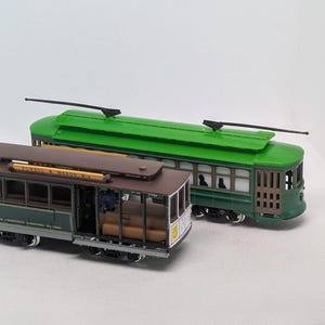 May include: Two model cable cars, one brown and one green, are displayed. The brown car has the text "Powell & Mason" and "San Francisco Municipal Railway" on it. The green car has a green roof and a similar design. Both have black poles.