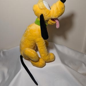 Puede incluir: Un peluche amarillo de Pluto con orejas y cola negras, un collar verde y una lengua rosa. El juguete est&aacute; sobre una superficie blanca.
