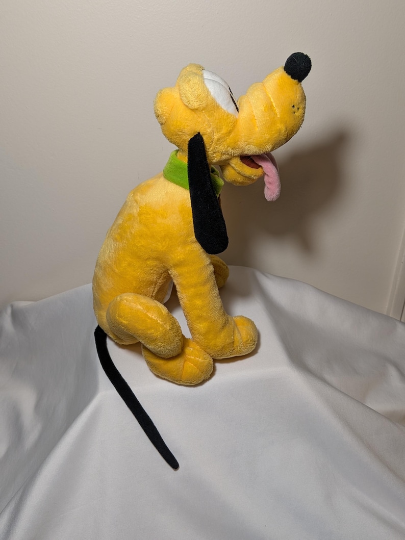 Puede incluir: Un peluche amarillo de Pluto con orejas y cola negras, un collar verde y una lengua rosa. El juguete est&aacute; sobre una superficie blanca.