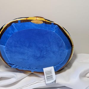 May include: A plush toy in the shape of a barrel, featuring a blue center and a yellow and brown rim. The toy has a soft, velvety texture and a tag with a barcode.