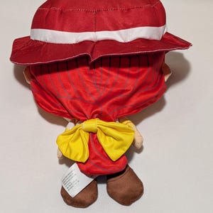 May include: A plush doll wearing a red hat with a white band and a red outfit, complemented by a yellow bow tie. The doll has brown shoes and a tag attached to one foot.