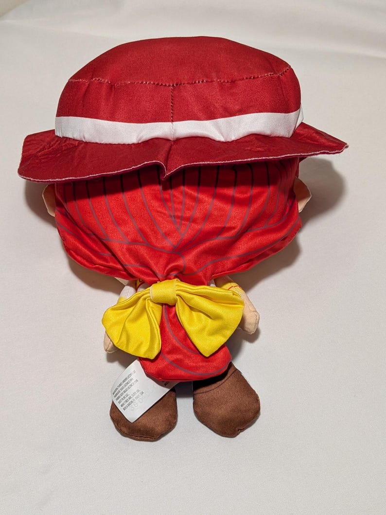 May include: A plush doll wearing a red hat with a white band and a red outfit, complemented by a yellow bow tie. The doll has brown shoes and a tag attached to one foot.