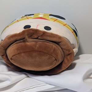 May include: A plush toy shaped like a cartoon character with a brown mustache and hat. The toy has black eyes, a red nose, and a yellow and white hat. The toy is resting on a white surface.