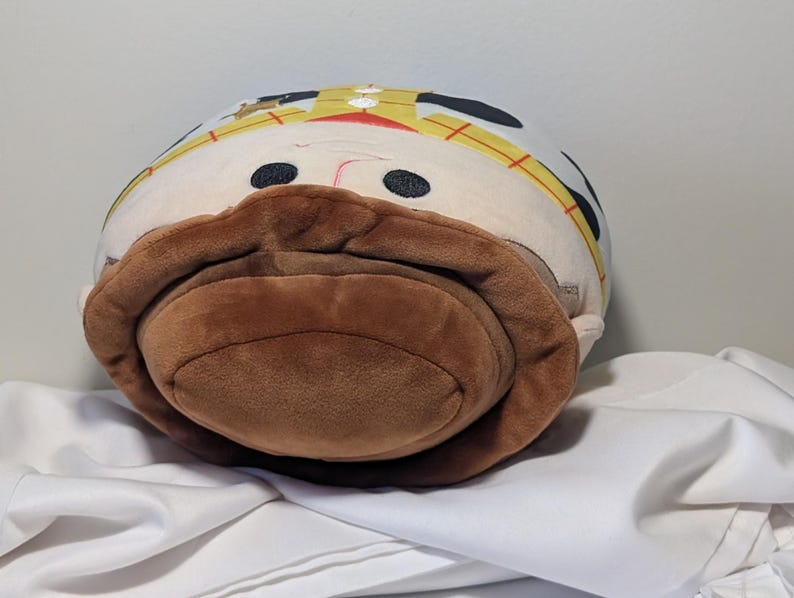 May include: A plush toy shaped like a cartoon character with a brown mustache and hat. The toy has black eyes, a red nose, and a yellow and white hat. The toy is resting on a white surface.