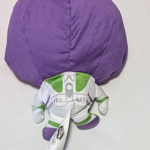 May include: A plush toy of Buzz Lightyear from Toy Story, featuring a large purple head and a white and green spacesuit. The toy has a tag with the Disney logo.