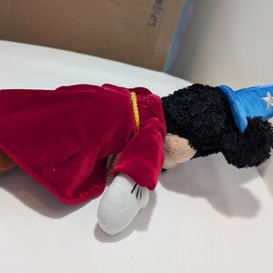 May include: A plush toy of a cartoon character dressed as a sorcerer. The toy has black fur, a blue hat with a white star, a red velvet cape, and white gloves. The toy is lying on a white surface.
