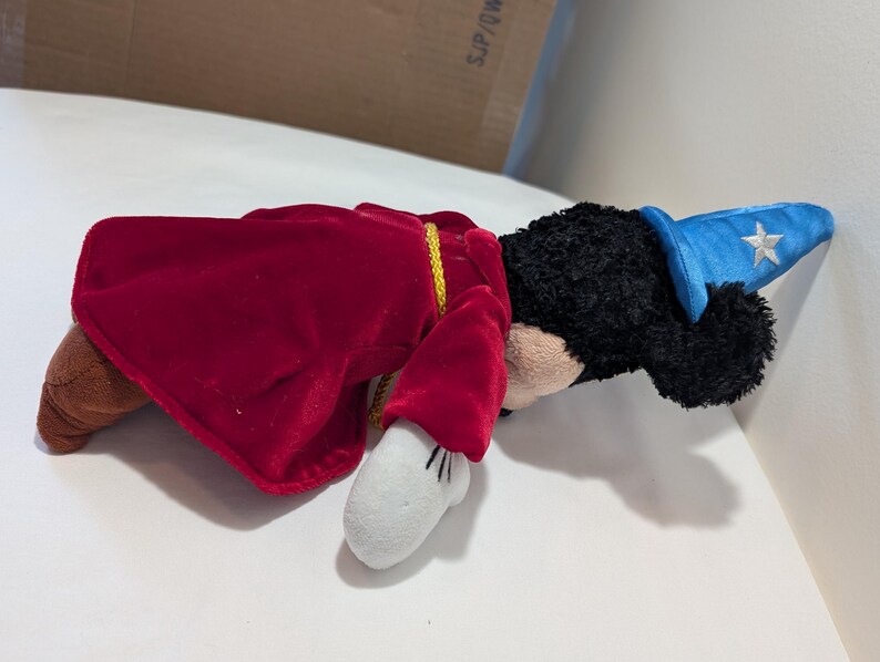 May include: A plush toy of a cartoon character dressed as a sorcerer. The toy has black fur, a blue hat with a white star, a red velvet cape, and white gloves. The toy is lying on a white surface.