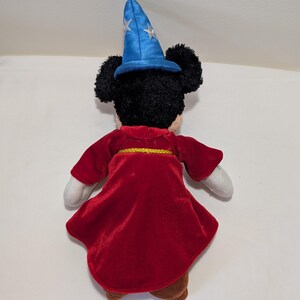 May include: A plush Mickey Mouse toy dressed as a sorcerer, viewed from the back. The toy wears a red velvet robe with a gold trim and a blue wizard hat with silver stars. The toy has black fur, white gloves, and brown shoes.
