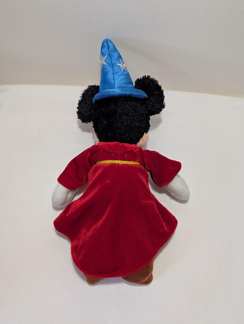 May include: A plush Mickey Mouse toy dressed as a sorcerer, viewed from the back. The toy wears a red velvet robe with a gold trim and a blue wizard hat with silver stars. The toy has black fur, white gloves, and brown shoes.