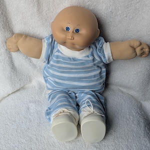 Puede incluir: Una muñeca calva con cabeza redonda de vinilo, ojos azules y cuerpo de tela suave. La muñeca viste una camiseta de manga corta a rayas azul claro y blanco con pantalones a juego, junto con zapatos blancos con cordones blancos atados.