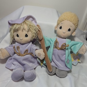 Könnte beinhalten: Zwei Plüschpuppen mit blonden Haaren und gestickten Gesichtern. Eine Puppe trägt ein lavendelfarbenes Outfit mit passendem Hut, die andere trägt ein lavendelfarbenes Outfit mit einem türkisfarbenen Umhang und hält einen Holzstab. Beide Puppen haben beige Seilakzente.