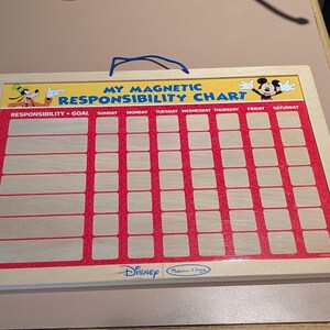 Op de afbeelding: Houten magnetisch verantwoordelijkheidsoverzicht met een rood en wit raster, met Disney-personages Goofy en Mickey Mouse. Het overzicht bevat de tekst "My Magnetic Responsibility Chart" en het Disney-logo.