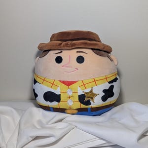 May include: A plush toy of a cartoon character, Sheriff Woody, from the movie Toy Story. The toy has a brown hat, a tan face with black eyes and a pink nose, and a yellow shirt with a red vest and a sheriff's badge.