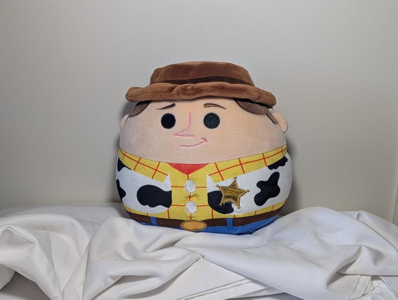 May include: A plush toy of a cartoon character, Sheriff Woody, from the movie Toy Story. The toy has a brown hat, a tan face with black eyes and a pink nose, and a yellow shirt with a red vest and a sheriff's badge.