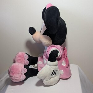 May include: A plush Minnie Mouse doll in a seated position. The doll features black ears, a pink bow, and a pink dress with white polka dots. The doll's hands are white with black stripes.
