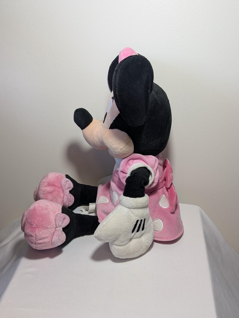 May include: A plush Minnie Mouse doll in a seated position. The doll features black ears, a pink bow, and a pink dress with white polka dots. The doll's hands are white with black stripes.