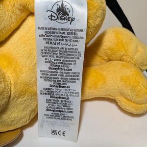 Puede incluir: Primer plano de un juguete de peluche amarillo con una etiqueta de Disney. La etiqueta incluye texto en varios idiomas, incluidos ingl&eacute;s, franc&eacute;s y espa&ntilde;ol. La etiqueta tambi&eacute;n incluye el logotipo de Disney e informaci&oacute;n sobre el origen y la distribuci&oacute;n del producto.