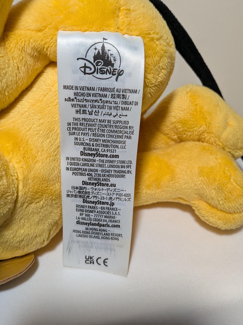 Puede incluir: Primer plano de un juguete de peluche amarillo con una etiqueta de Disney. La etiqueta incluye texto en varios idiomas, incluidos ingl&eacute;s, franc&eacute;s y espa&ntilde;ol. La etiqueta tambi&eacute;n incluye el logotipo de Disney e informaci&oacute;n sobre el origen y la distribuci&oacute;n del producto.