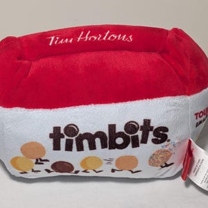 Puede incluir: Un peluche con forma de caja de Timbits Tim Hortons. La parte superior es roja, el centro es blanco con la palabra "timbits" e ilustraciones de Timbits. El logotipo de Tim Hortons está en la parte superior. Una etiqueta está adjunta.