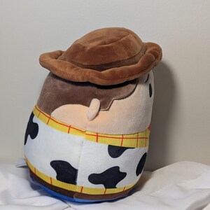 May include: A plush toy of the character Woody from the movie Toy Story. The toy is brown, tan, and white with black spots, and a brown cowboy hat. The toy has a blue base and yellow trim.